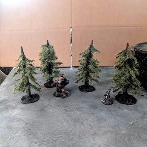 Può includere: Quattro modelli di pini in miniatura con fogliame verde testurizzato e tronchi marrone scuro, disposti accanto a due piccole statuette fantasy dipinte su una superficie grigia. Gli alberi sono di forma conica con dettagli dei rami.
