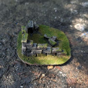 May include: A miniature diorama base featuring a weathered gray stone wall ruin partially covered in vibrant green moss. The structure is set on a textured, grass-covered platform, creating a realistic landscape element for tabletop gaming models.