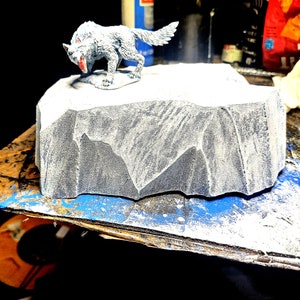 Puede incluir: Una figurita de criatura blanca similar a un lobo se encuentra sobre una base rocosa gris. La base es texturizada y parece estar hecha de un material rugoso.