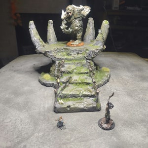 May include: A miniature stone altar with a green moss covering and a small, green, stone creature on top. The altar has a set of stairs leading up to the creature. Two miniature figures stand on the ground in front of the altar.