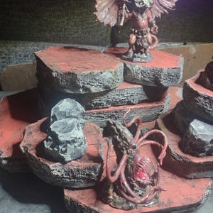 Puede incluir: Un conjunto de terreno de juego de mesa de varios niveles, pintado en rojo y gris, que parece roca. El conjunto incluye un demonio rojo con alas y una criatura con tentáculos.