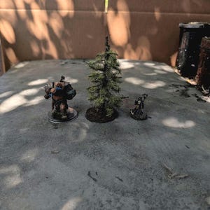 May include: A painted miniature figurine of a large, muscular creature with dark armour, a miniature model of a tall, thin evergreen tree with green foliage, and a small, painted miniature figurine of a robed figure holding a staff.