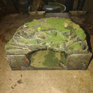 May include: A miniature, moss-covered rock formation with a cave opening. The model features a layered stone design with green vegetation on top and inside the cave. The color palette includes shades of gray, brown, and green.