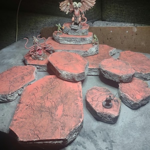 Puede incluir: Un conjunto de piezas de terreno para juegos de mesa de forma irregular, con aspecto de roca, pintadas en rojo y gris. Las piezas están dispuestas en forma circular, con una pieza más grande en el centro. Una figura en miniatura de un demonio con alas está de pie sobre la pieza central.