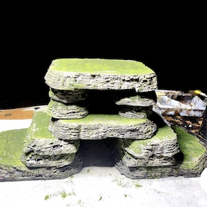 Puede incluir: Una pila de estructuras rocosas grises y verdes con una abertura en forma de cueva en el medio. Las estructuras están cubiertas de musgo verde.