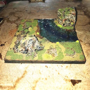 May include: A small-scale diorama depicting a river scene with rocky outcrops and grassy banks. The river is a deep blue, and the rocks are textured. Green and yellow vegetation is spread across the landscape, creating a naturalistic effect.
