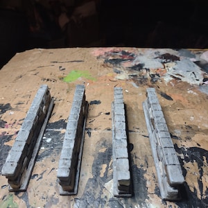 May include: Four miniature stone wall sections, each with a rough, textured surface, painted in a light gray color. The walls are arranged in a row on a brown surface.