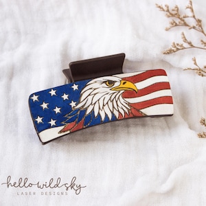 Może przedstawiać: Prostokątna spinka do włosów z patriotycznym wzorem. Spinka przedstawia amerykańską flagę z ilustracją głowy orła. Projekt wykorzystuje kolory czerwony, biały i niebieski, z tekstem "hello wild sky laser designs".