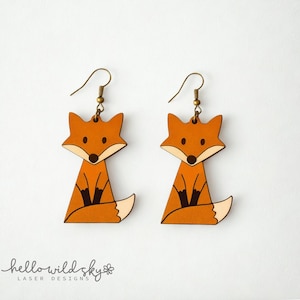 May include: A pair of wooden fox earrings. Each earring features a stylized fox design with orange, white, and black details. The earrings have gold-colored hooks. The text "hello wild sky" is at the bottom.