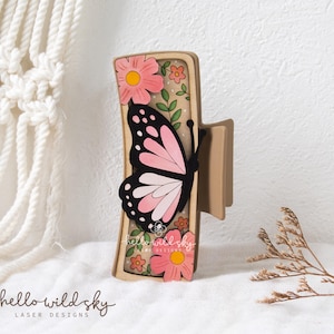 May include: A tan hair claw clip featuring a layered design with a large pink and black butterfly, pink flowers, and green leaves. The surface includes a light tan background with white dots and the text hello wild sky laser designs.