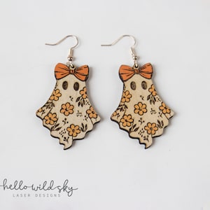 May include: A pair of wooden ghost earrings with orange bows and floral patterns. The earrings have silver hooks and are laser cut. The ghost-shaped earrings are decorated with orange flowers and leaves. The earrings are a digital download from Hello Wild Sky Laser Designs.