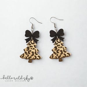 May include: A pair of Christmas tree earrings with a leopard print design. Each earring features a black bow above the tree shape and a silver hook. The earrings are made of wood and are a digital download.