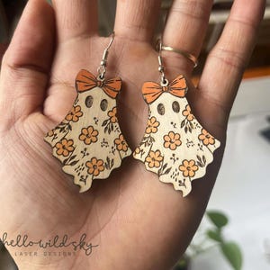 May include: Wooden ghost earrings with orange bows and floral patterns. The earrings have a laser-cut design with a light wood finish. The text "hello wild sky" is visible in the image.