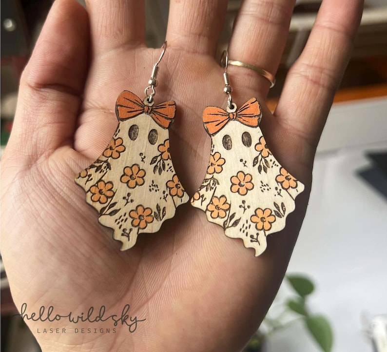 May include: Wooden ghost earrings with orange bows and floral patterns. The earrings have a laser-cut design with a light wood finish. The text "hello wild sky" is visible in the image.