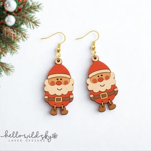 Può includere: Un paio di orecchini in legno a forma di Babbo Natale. Ogni orecchino ha un cappello rosso, una barba beige e un abito marrone con fibbia dorata. Gli orecchini pendono da fili a gancio dorati. Lo sfondo è bianco con un rametto di sempreverde.