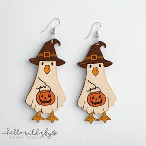 May include: A pair of wooden Halloween earrings. Each earring features a light-colored chicken-like figure wearing a brown witch hat with a buckle. The chicken holds an orange jack-o'-lantern. The earrings have silver hooks.