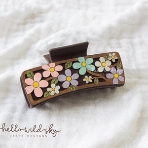 May include: Brown floral hair claw clip with painted pink, blue, and purple flowers. The clip has a rectangular design with a dark brown base and a spring-loaded mechanism. The text "hello wild sky" is visible.