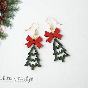 Puede incluir: Un par de pendientes de árbol de Navidad con lazos rojos y formas de árbol verdes. Los pendientes tienen ganchos dorados. La imagen también incluye el texto "hello wild sky" y "laser designs".