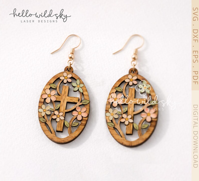 May include: Wooden Easter egg earrings with a cross design, surrounded by flowers in shades of pink, green, and cream. The earrings have gold-colored hooks and the text "hello wild sky laser designs".