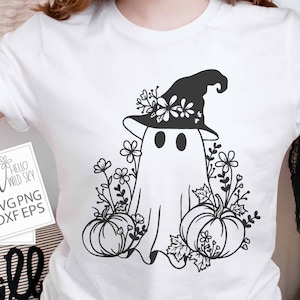 May include: A white t-shirt with a black and white graphic of a ghost wearing a witch hat and holding a pumpkin. The ghost is surrounded by flowers and leaves. The text "Hello Wild Sky" is on a white tag.