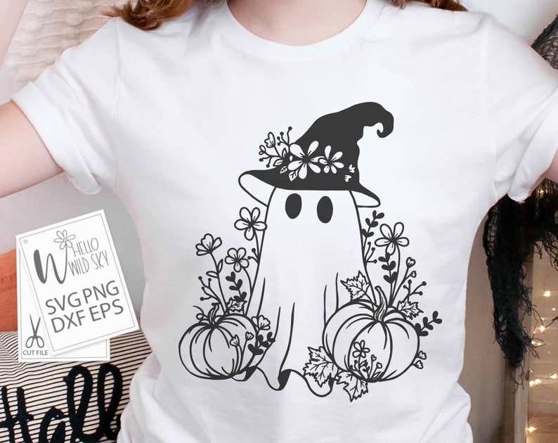 May include: A white t-shirt with a black and white graphic of a ghost wearing a witch hat and holding a pumpkin. The ghost is surrounded by flowers and leaves. The text "Hello Wild Sky" is on a white tag.