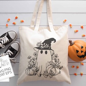 May include: A white tote bag with a black and white design of a ghost wearing a witch hat and surrounded by flowers and pumpkins. The text "Hello Wild Sky" is on a white card in the lower left corner of the image.