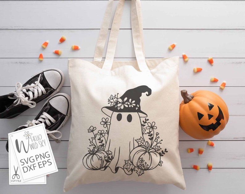 May include: A white tote bag with a black and white design of a ghost wearing a witch hat and surrounded by flowers and pumpkins. The text "Hello Wild Sky" is on a white card in the lower left corner of the image.