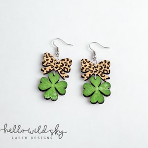 May include: Wooden earrings featuring a green shamrock design with a leopard print bow. The earrings have silver-colored hooks. The words "hello wild sky" and "laser designs" are printed at the bottom of the image.