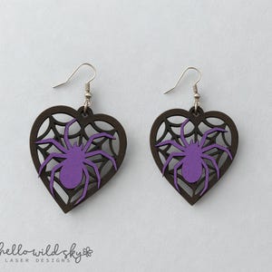 May include: A pair of heart-shaped earrings with a spider and web design. The earrings feature a dark brown heart frame with a purple spider in the centre. The spider is set against a web design. The earrings have silver-coloured hooks.