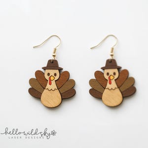 May include: A pair of wooden turkey earrings with gold-colored hooks. Each earring features a turkey design with a brown hat, a light brown body, and multi-toned brown feathers. The earrings are a digital download.