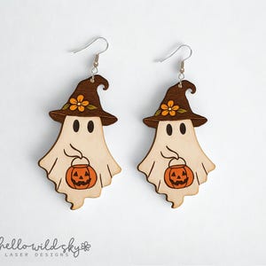 May include: Two wooden Halloween ghost earrings. Each ghost wears a brown witch hat with an orange flower and holds an orange jack-o'-lantern. The earrings are laser cut and have silver earring hooks. The brand name "hello wild sky" is at the bottom.