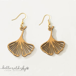 May include: Two wooden ginkgo leaf earrings with gold-coloured ear wires. The earrings are light brown and laser cut with detailed lines. The words "hello wild sky" are at the bottom. The earrings are a delicate design.