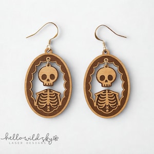 Könnte beinhalten: Ein Paar Holz-Skelett-Ohrringe. Jeder Ohrring hat ein Schädel- und Brustkorb-Design in einem ovalen Rahmen. Die Ohrringe sind hellbraun mit goldfarbenen Ohrhaken. Der Text "hello wild sky laser designs" befindet sich unten.