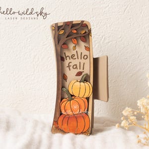 May include: A layered wooden hair claw clip featuring a laser-cut design of a tree with falling leaves, three orange pumpkins, and the text hello fall. The design includes brown, tan, and orange tones, showcasing a detailed autumn-themed illustration.