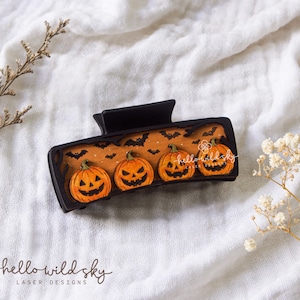 May include: A black rectangular hair claw clip featuring an orange inlay with four carved jack-o'-lantern pumpkins and black bat silhouettes. The text "hello wild sky" is visible on the clip, and "SVG . DXF . EPS . PDF DIGITAL DOWNLOAD" is on the side.