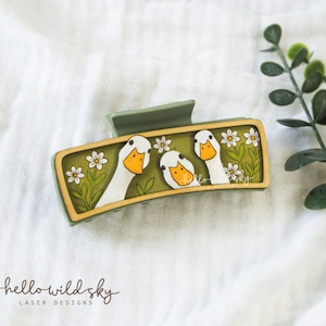 May include: A rectangular sage green hair claw clip featuring a printed design of three white geese with orange beaks surrounded by white daisies on a green background. The clip has a light wood-toned border and a sturdy spring mechanism.