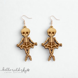 May include: Two wooden skeleton ballerina earrings with gold-colored hooks. The earrings feature a skeleton head, rib cage, and tutu design. The earrings are laser cut and have a light brown color. The words "hello wild sky" are at the bottom.
