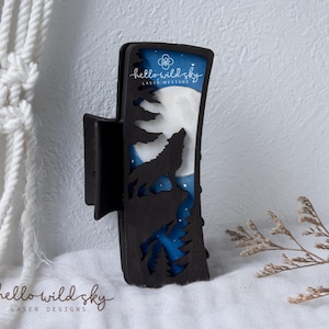 May include: A black hair claw clip featuring a laser-cut design of a wolf howling at a full moon against a blue night sky with pine trees. The clip displays the text hello wild sky LASER DESIGNS and the side text reads SVG. DXF. EPS. PDF DIGITAL DOWNLOAD.