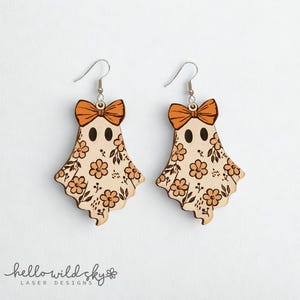 May include: A pair of wooden ghost earrings with orange bows and floral patterns. The earrings have silver hooks and are designed with two black eye dots. The earrings are a digital download from Hello Wild Sky Laser Designs.