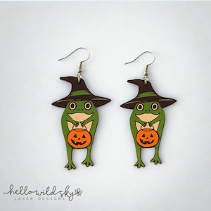May include: A pair of Halloween-themed earrings featuring green frog characters wearing witch hats and holding orange jack-o'-lanterns. The earrings are made of wood and have silver-colored ear wires. The design is a digital download from Hello Wild Sky Laser Designs.