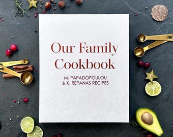 May include: A white cookbook with the title "Our Family Cookbook" in maroon text. The subtitle is "M. PAPADOPOULOU & K. REPANAS RECIPES" also in maroon text. The cookbook is surrounded by various food items, including limes, avocados, cranberries, and cinnamon sticks.