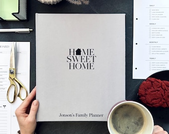 Könnte beinhalten: Ein hellgrauer Familienplaner mit dem Text "Home Sweet Home" und "Jonson's Family Planner" in Schwarz. Der Planer ist geöffnet, und eine Hand hält ihn. Weitere Artikel sind eine Schere, ein Stift und eine Tasse Kaffee.
