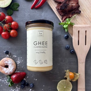 May include: A glass jar of ghee, a clarified butter, with a white label that reads "FOOD from LOVE GHEE CLARIFIED BUTTER tasty & healthy". The jar is on a gray surface with various fruits and vegetables around it.