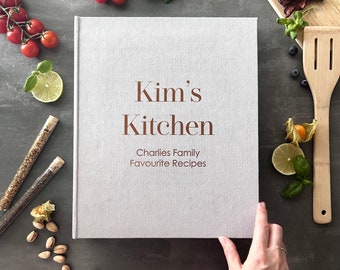 May include: A gray linen-covered cookbook with the title "Kim's Kitchen" and the subtitle "Charles Family Favourite Recipes".
