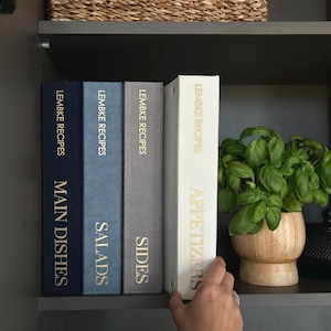 Op de afbeelding: Vier receptmappen in marineblauw, blauw, grijs en wit, met de labels "Hoofdgerechten", "Salades", "Bijgerechten" en "Voorgerechten" met gouden letters. Een basilicumplant in een houten pot staat ernaast.