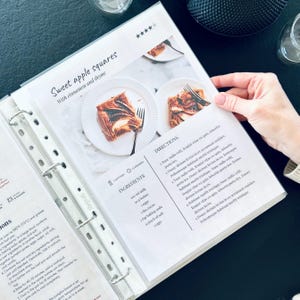 Può includere: Un raccoglitore di ricette aperto su una pagina con una ricetta per quadrati di mele dolci. La pagina presenta una foto del dolce finito su un piatto bianco con una forchetta. Il titolo della ricetta è in un carattere decorativo.