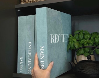 May include: Three teal recipe binders labeled "Baking", "Entertaining", and "Main Dishes" are displayed on a shelf. The largest binder reads "RECIPES" in white text. A hand is holding the "Main Dishes" binder. A black teapot and basil plant are also visible.