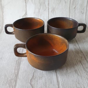 Puede incluir: Tres tazas de cerámica marrón con un esmalte marrón oscuro y un interior naranja. Las tazas tienen una forma redondeada y un asa en un lado.
