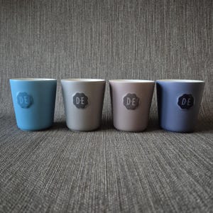 May include: Four ceramic cups in shades of blue, grey, and mauve are arranged in a row. Each cup has a small, embossed emblem with the letters "DE". The cups have a smooth, matte finish and a simple, modern design.