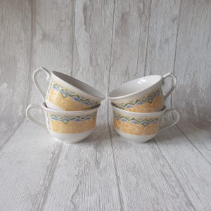 May include: Four white ceramic teacups with a yellow, blue, and green geometric pattern. The cups are stacked on top of each other.